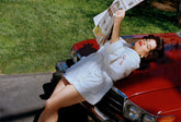 woman in white robe leaning against red car while reading a newspaper