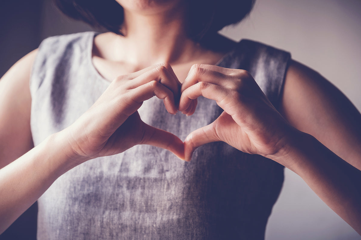 A close-up lifestyle shot of a woman wearing a textured gray sleeveless top, forming a heart shape with her hands against a soft, blurred background.