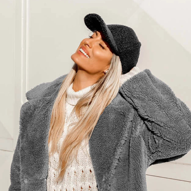 Woman wearing a gray coat, white sweater, and black hat against a light background