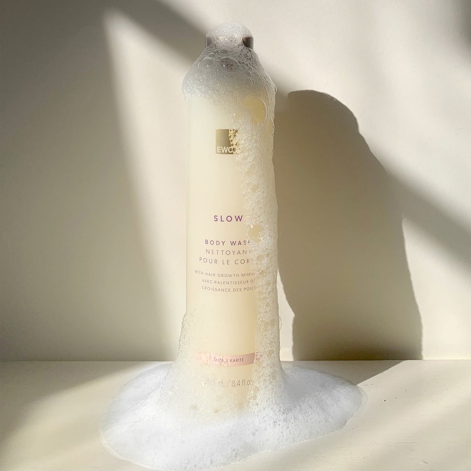 Bottle of EWC Slow Shea Body Wash with soap suds on a light background