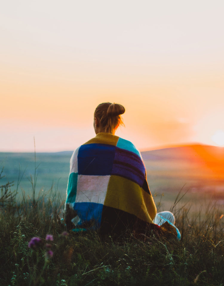 A person sitting on a grassy hill at sunset, seen from behind, wrapped in a multicolor patchwork blanket. The background features a warm orange and yellow sunset over a distant landscape.