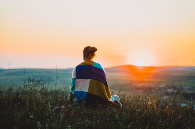 A person sitting on a grassy hill at sunset, seen from behind, wrapped in a multicolor patchwork blanket. The background features a warm orange and yellow sunset over a distant landscape.