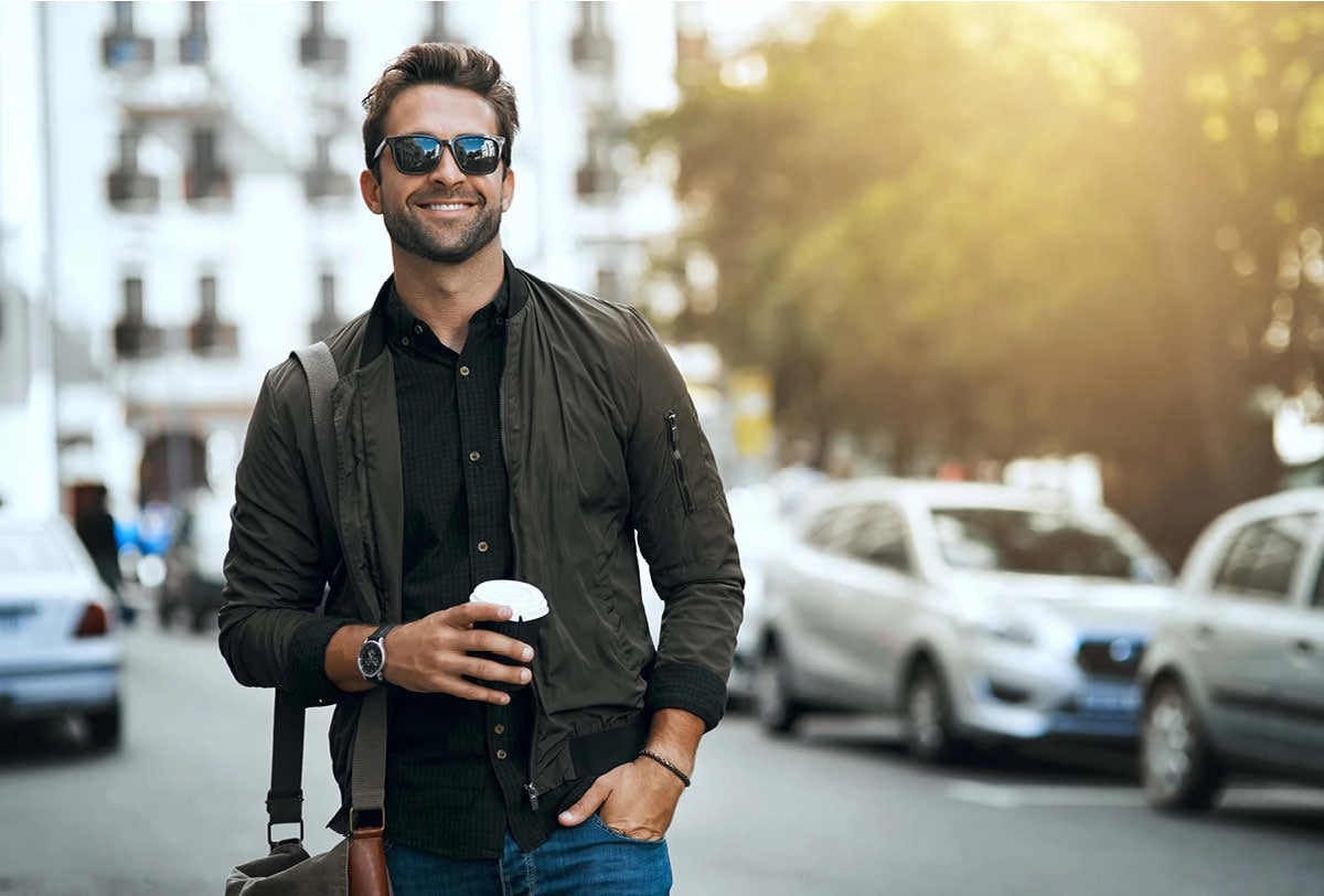 Man walking on a city street holding a coffee cup, wearing sunglasses and a jacket.