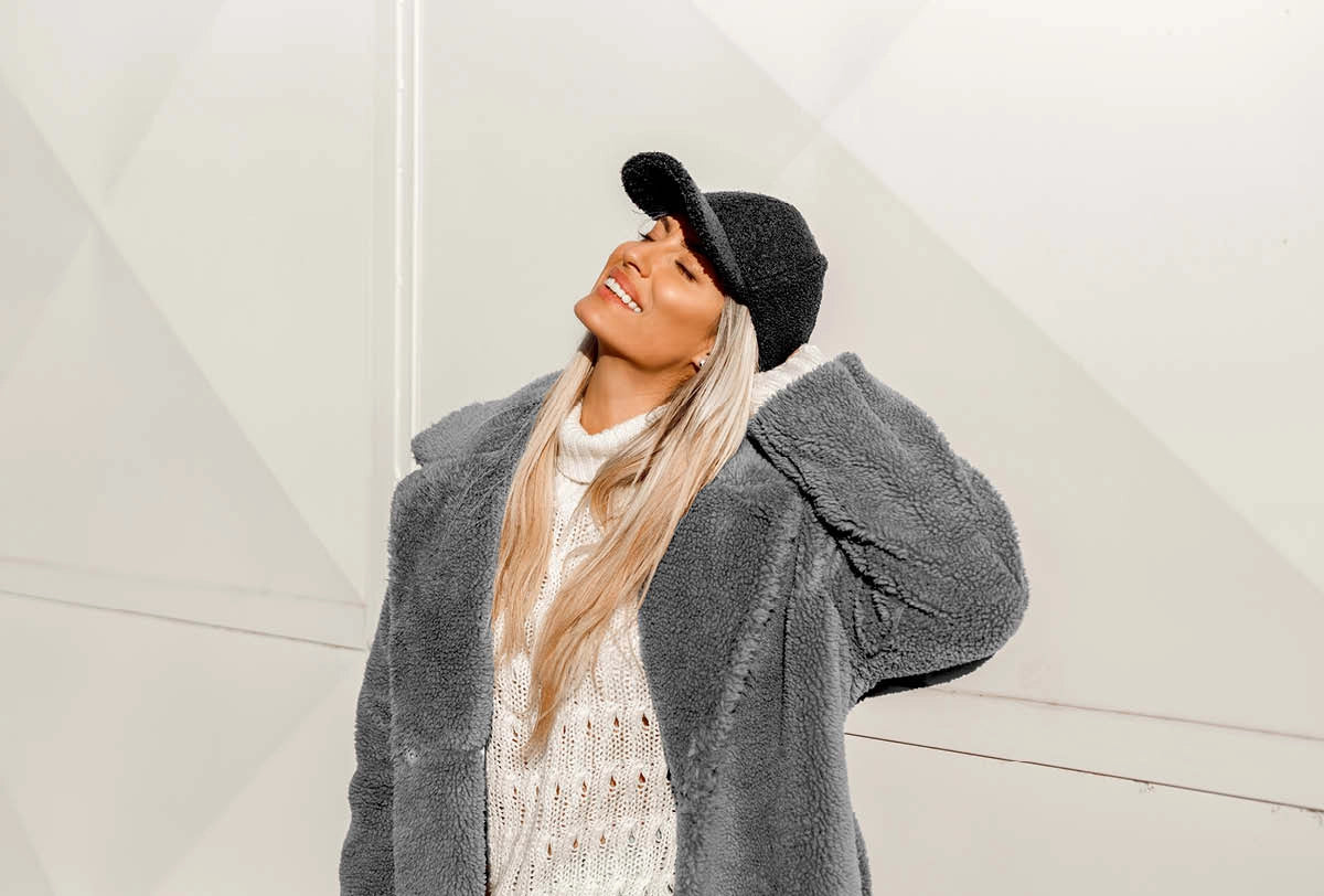Woman wearing a gray coat, white sweater, and black hat against a light background