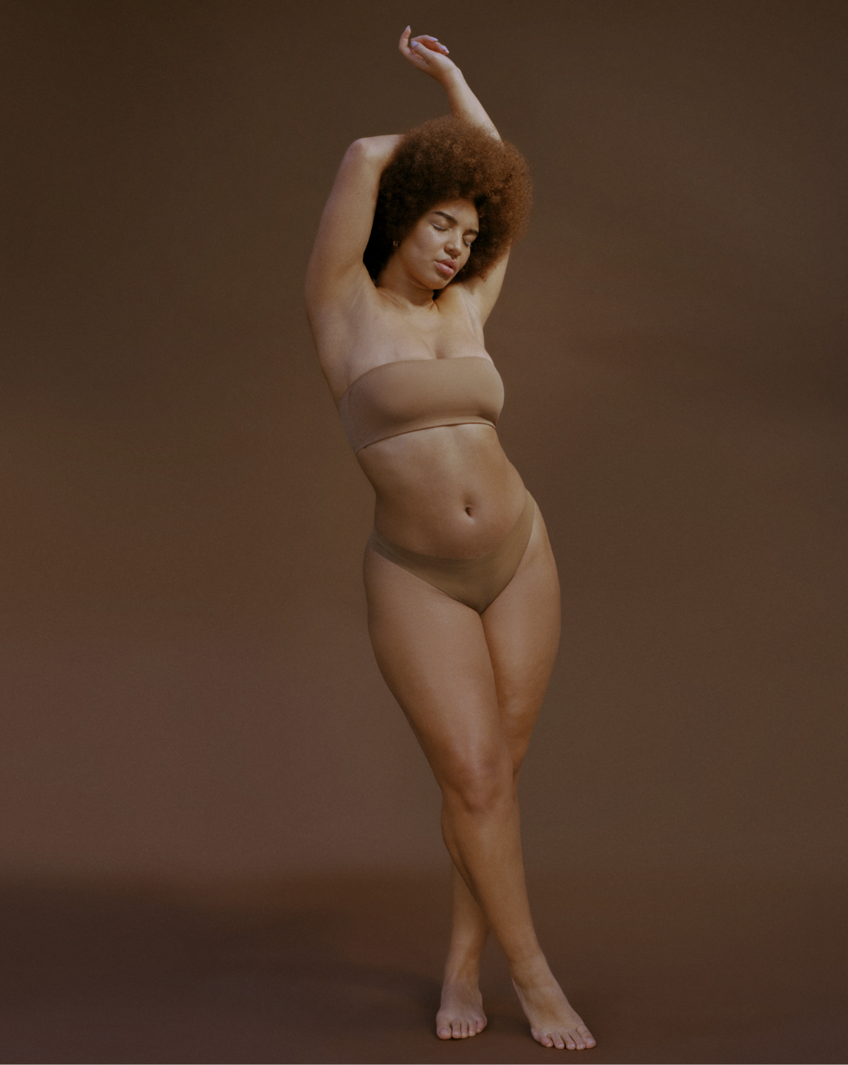 standing woman with arms crossed over her head wearing nude colored bra and underwear