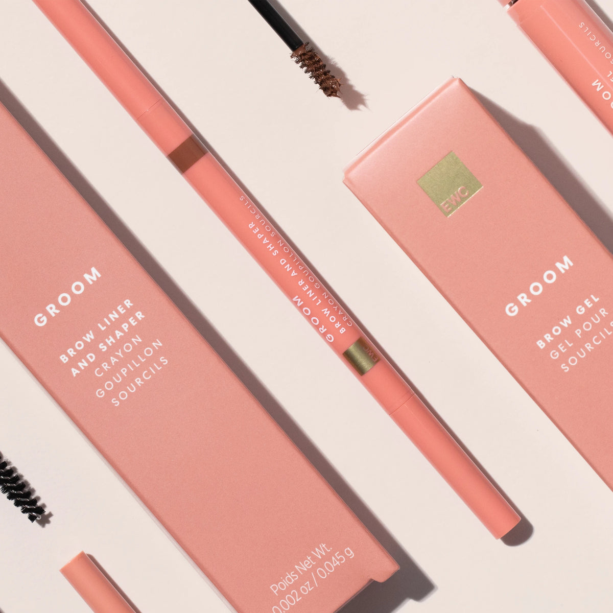 A flat-lay product shot of a Groom eyebrow product set, featuring a peach-pink brow pencil, matching brow gel stick, and their corresponding pink packaging boxes on a light beige background.