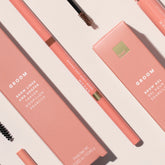 A flat-lay product shot of a Groom eyebrow product set, featuring a peach-pink brow pencil, matching brow gel stick, and their corresponding pink packaging boxes on a light beige background.