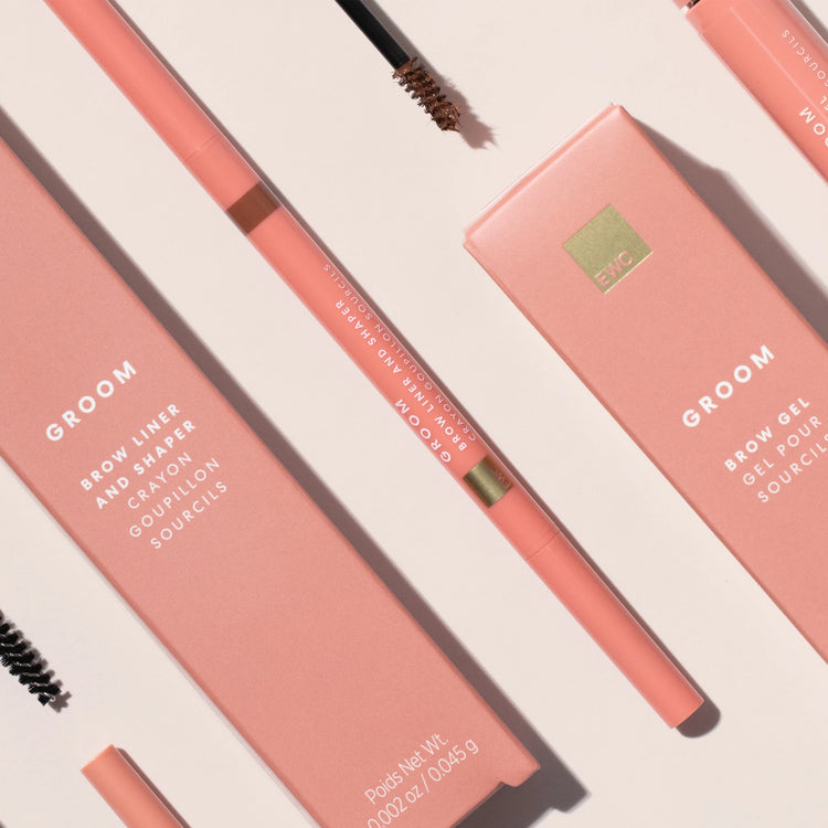 A flat-lay product shot of a Groom eyebrow product set, featuring a peach-pink brow pencil, matching brow gel stick, and their corresponding pink packaging boxes on a light beige background.