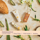 Travel-sized EWC SLOW® Summer Oasis collection products (Body Wash, Body Polish, and Shimmer Body Oil) arranged over a light stone surface. Products are surrounded with natural elements (coconut, bamboo stems, orchids and aloe leaves). The text reads "Fragrance notes of coconut water, ylang-ylang, wild orchid and bamboo." 