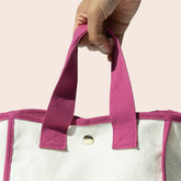 White tote bag with berry colored handles held by a hand against a light pink background
